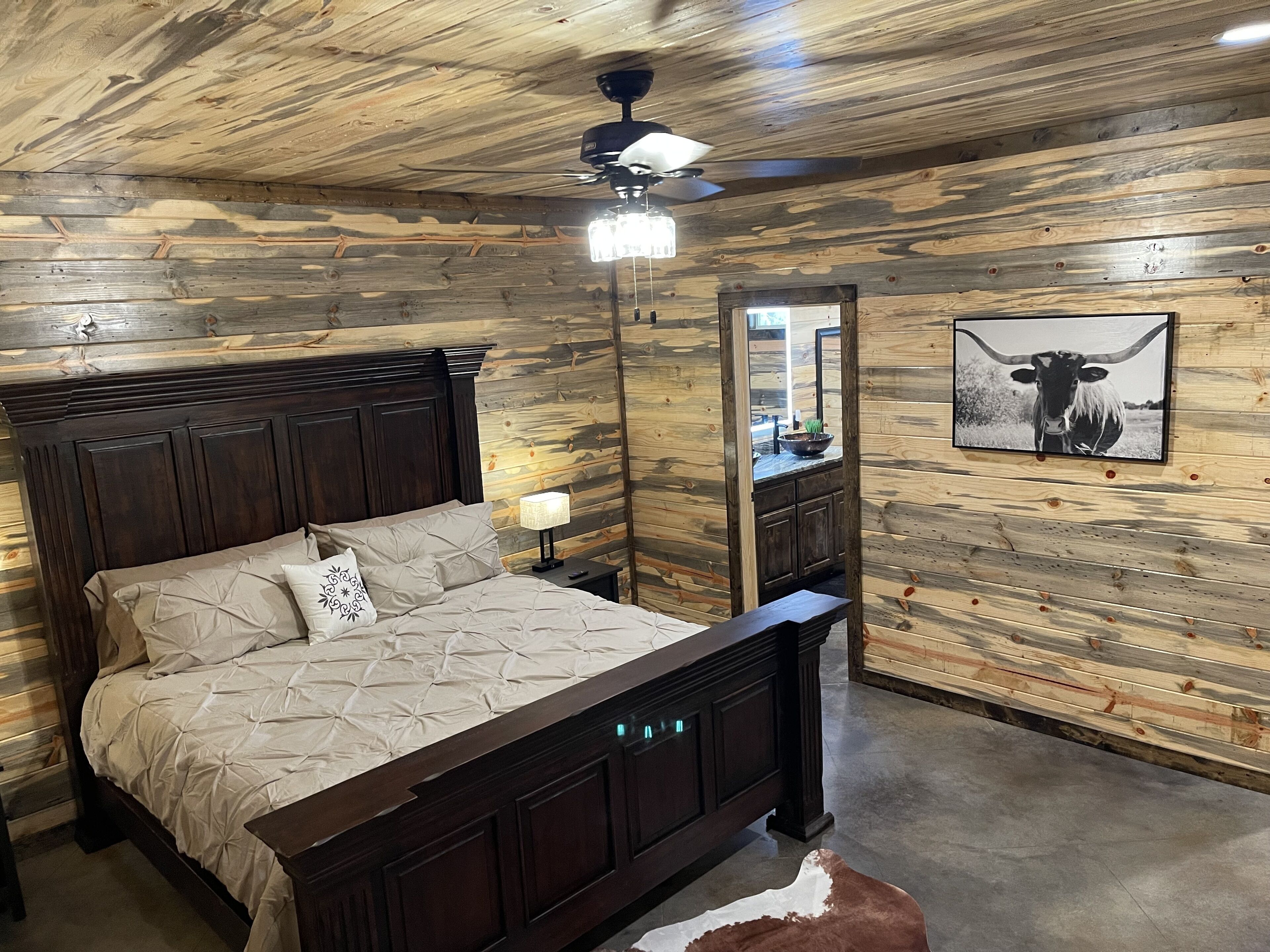 A bedroom with a wooden ceiling, a bed with a headboard, a nightstand, a lamp, a mirror, and a cow picture on the wall.