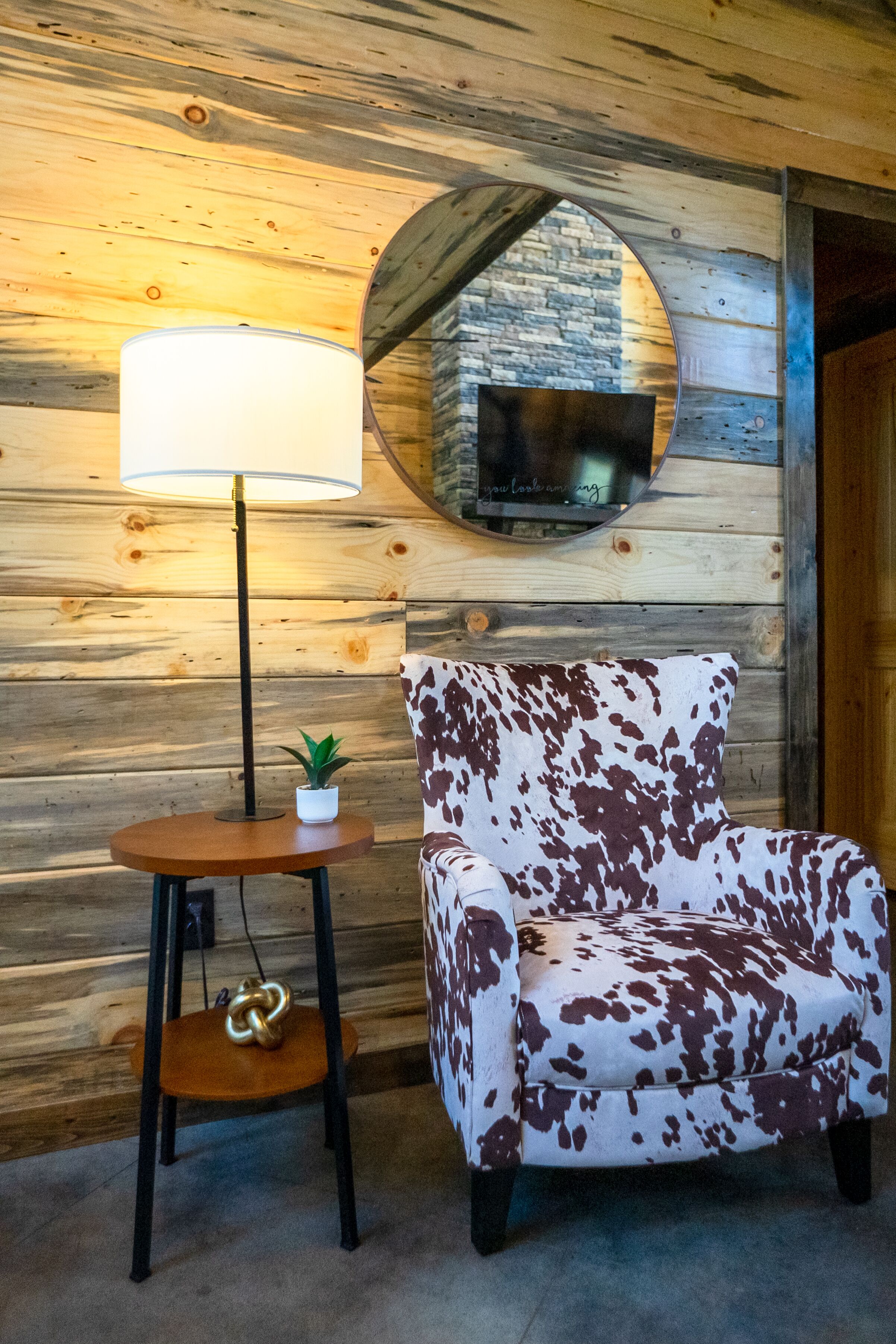 A cozy room with a wooden wall, a round mirror, a leather chair, a small table with a lamp, and a potted plant.