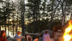 Eine Gruppe von Menschen sitzt an einem Lagerfeuer in einem bewaldeten Gebiet mit Holzmöbeln und einem Steintisch.