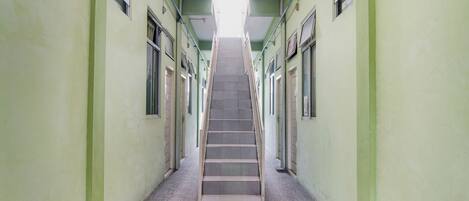 A long, narrow hallway with a central staircase leading to an upper floor. The walls are painted a light green, and there are windows on both sides of the hallway.