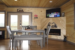 Un intérieur en bois comprenant une table à manger, des chaises, une cheminée et un téléviseur fixé au mur, diffusant des services de streaming.