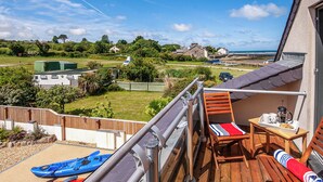 Un balcon avec des kayaks, une table sur laquelle se trouve un plateau rempli d’objets, et une vue sur une zone côtière avec des maisons et de la végétation.