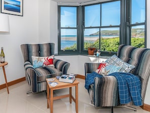 A room with two striped armchairs, a small wooden table, and a view of the sea through large windows.