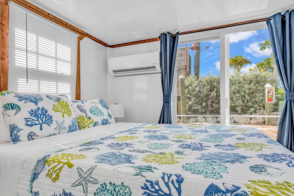 A bedroom with a bed, a window with blinds, an air conditioning unit, and a view of the outdoors.