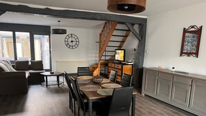 Un coin repas avec une table en bois, des chaises noires et des sets de table en fibres tressées. Une horloge et un téléviseur sont fixés au mur.
