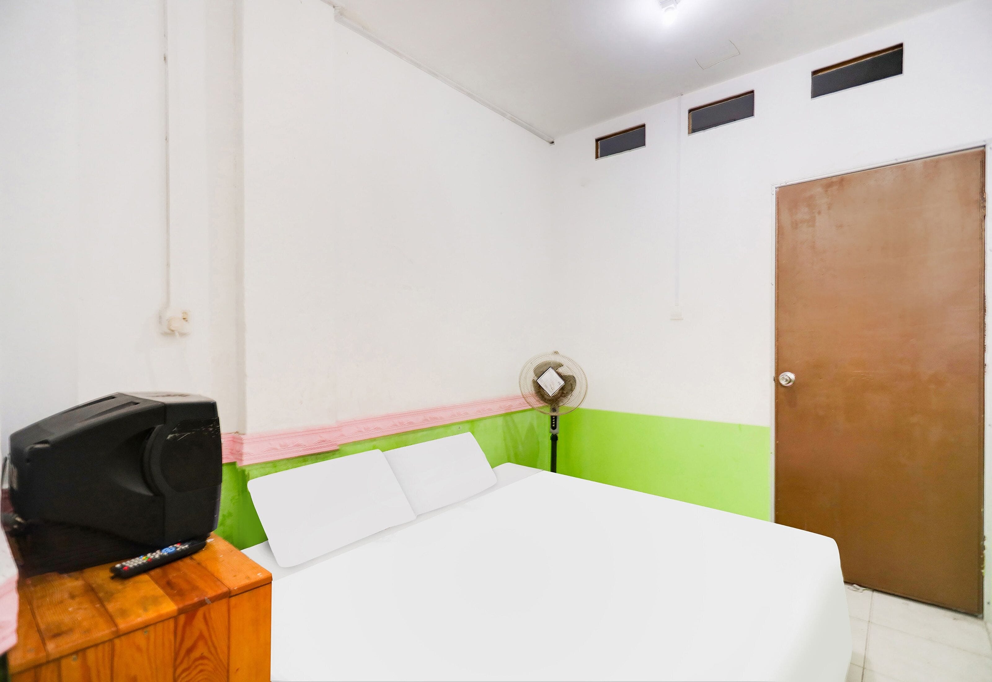 A room with a bed, a desk with a remote control, a wall-mounted fan, and a door.