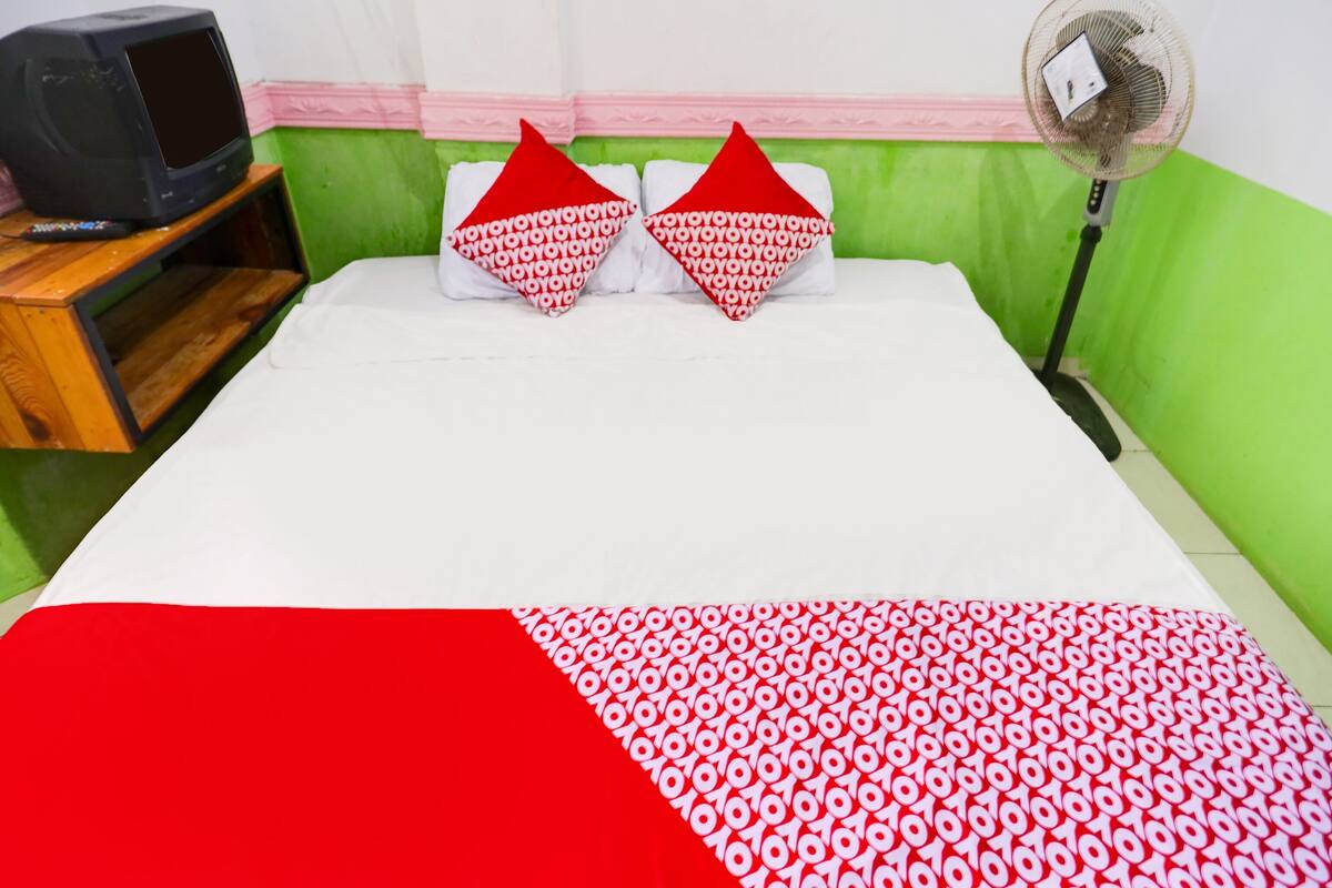Un lit recouvert d’une couvre-lit à motifs rouges et blancs, deux oreillers en forme de cœur aux motifs rouges et blancs, un téléviseur posé sur un meuble en bois et un ventilateur sur pied.