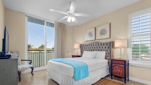 A bedroom with a large bed, a ceiling fan, a window with blinds, and a view of palm trees.