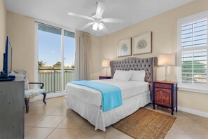 A bedroom with a large bed, a ceiling fan, a window with blinds, and a view of palm trees.