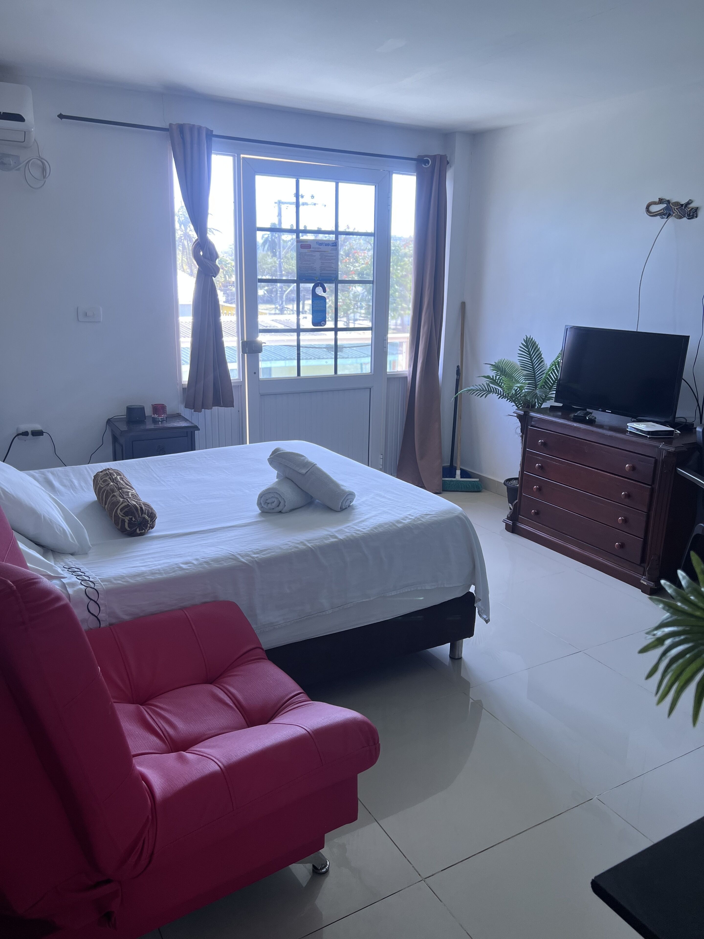 Une chambre d’hôtel avec un lit, un fauteuil rouge, un téléviseur posé sur une commode et une fenêtre avec des rideaux.