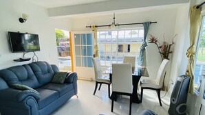 Un salon avec un canapé bleu, une table à manger avec des chaises, un téléviseur fixé au mur et une grande fenêtre avec des rideaux.