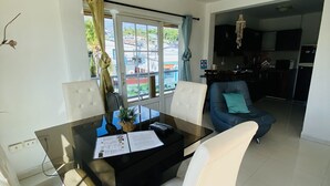 Un salon moderne avec une table à manger en verre, des chaises à revêtement blanc, un canapé bleu et une vue sur l’extérieur grâce à une grande fenêtre.