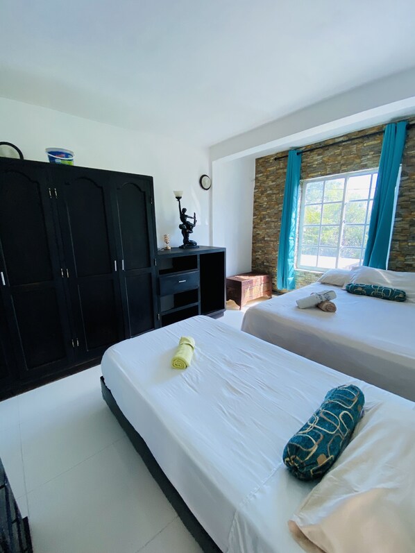 A hotel room with two beds, a wardrobe, a desk with a lamp, a window with curtains, and a stone wall.