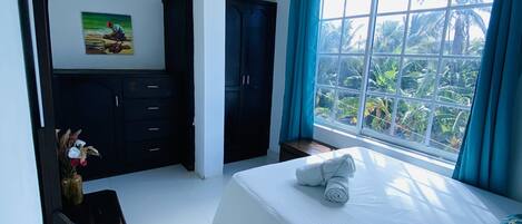 A modern hotel room with a large bed, a wardrobe, a desk, and a window with a view of palm trees.