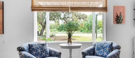 A room with two blue armchairs with star patterns, a small round table, a window with a bamboo blind, and a view of greenery outside.