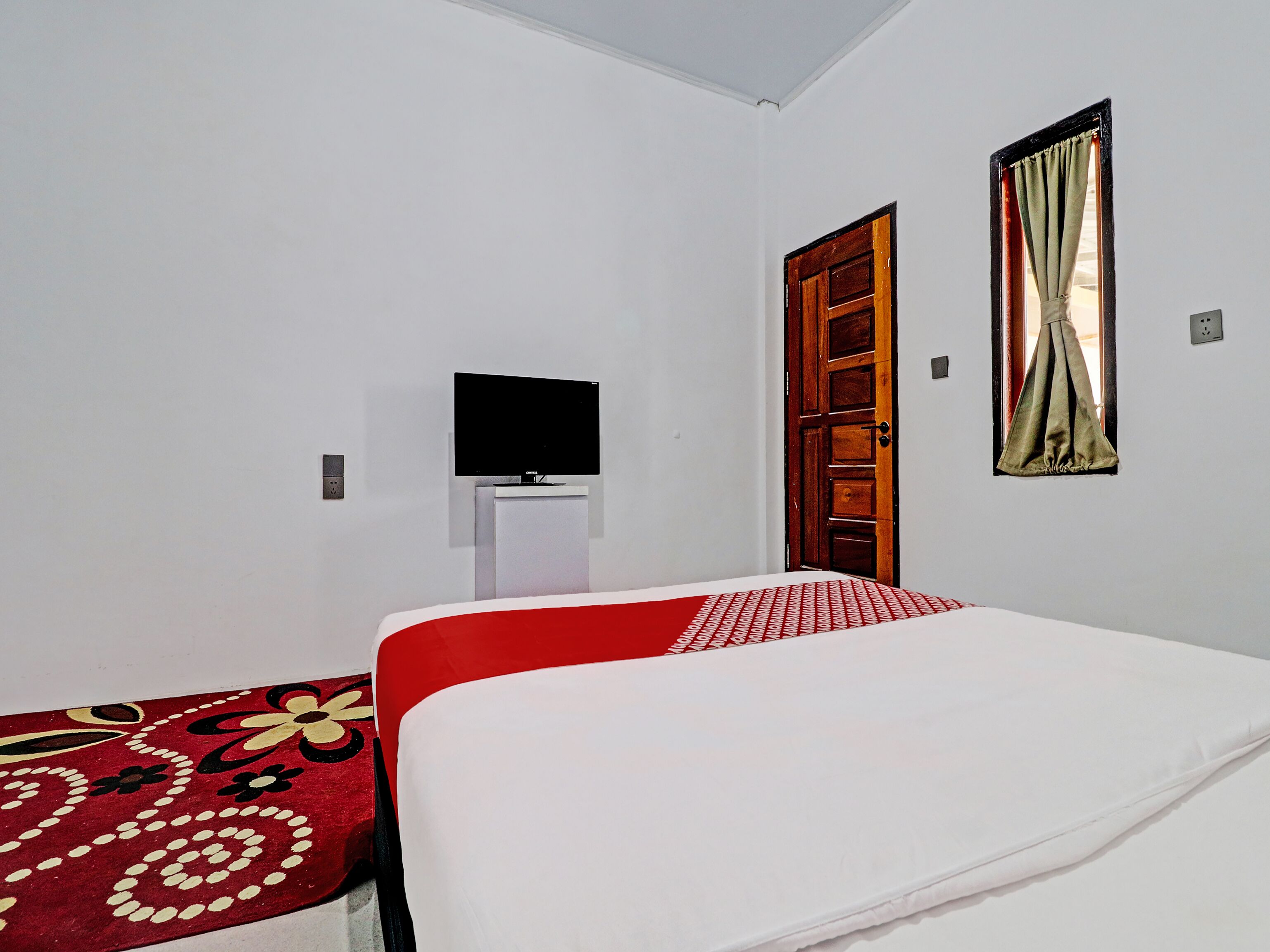 Una habitación con una cama, un televisor sobre un mueble, una puerta y una ventana con cortinas.
