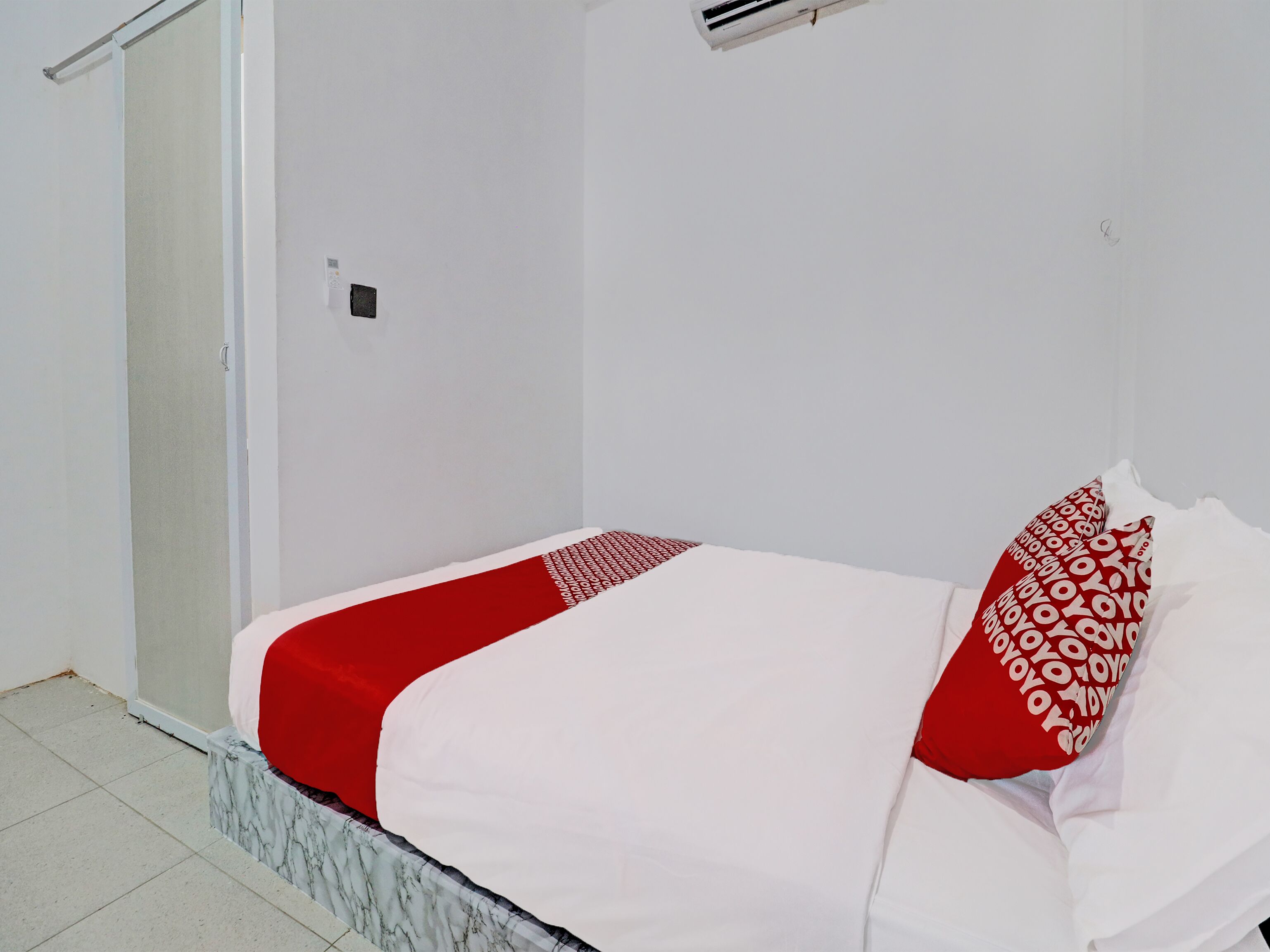 Una cama pequeña y bien arreglada con colcha y almohadas blancas y rojas, una mesita de noche de mármol y una pared blanca con un pequeño equipo de aire acondicionado.