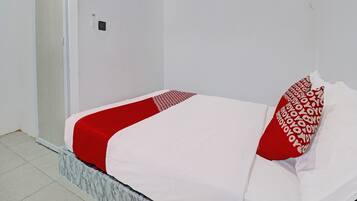 Una cama pequeña y bien arreglada con colcha y almohadas blancas y rojas, una mesita de noche de mármol y una pared blanca con un pequeño equipo de aire acondicionado.