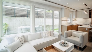 Een moderne woonkamer met een bank, fauteuil en salontafel. Er is een keuken met kasten en een eettafel. Grote ramen zorgen voor natuurlijk licht en uitzicht op de achtertuin.
