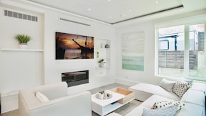 Een moderne woonkamer met een bank, een salontafel en een open haard. Daarboven hangt een groot schilderij en er is een raam met uitzicht op buiten.