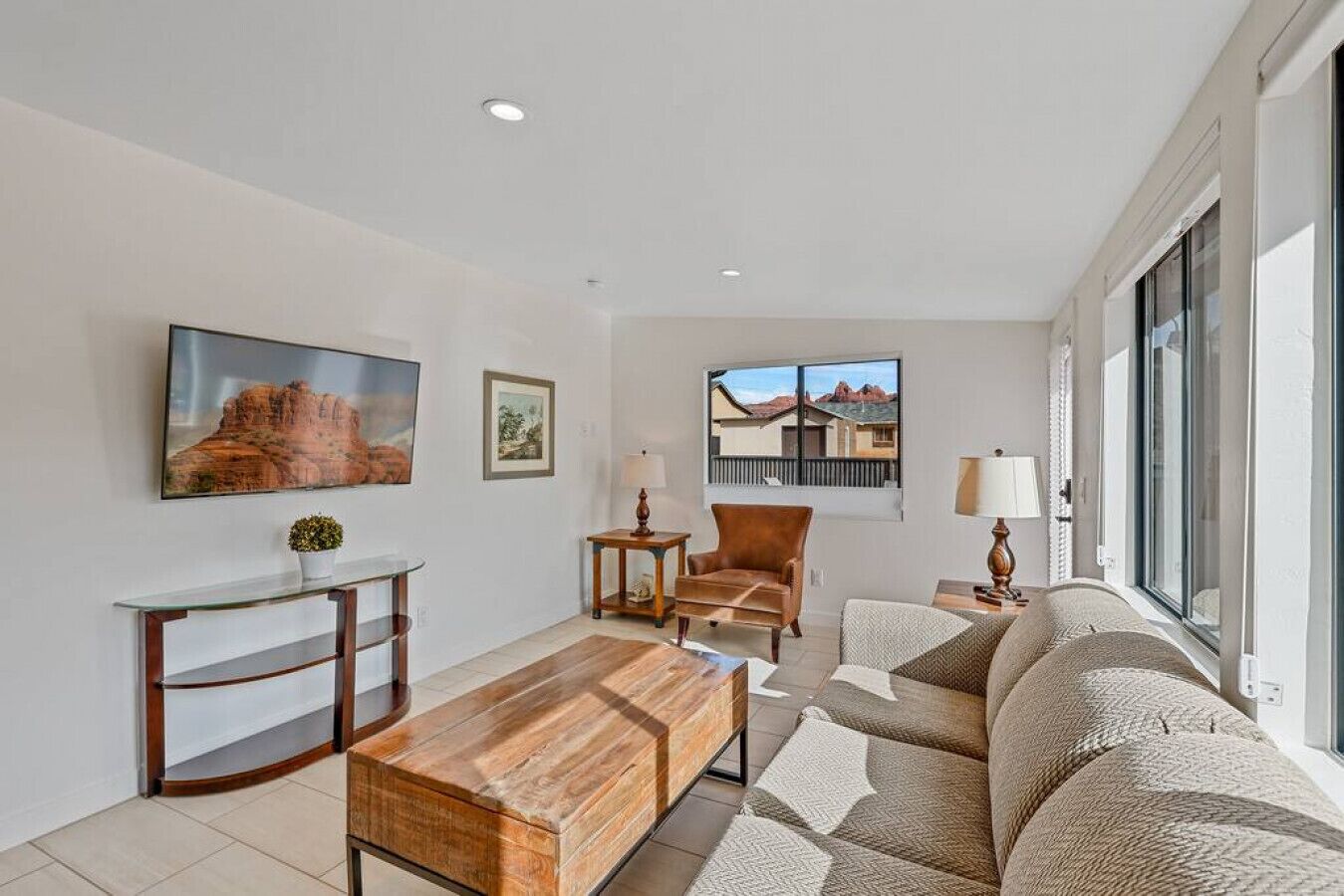 A modern living room with a wooden coffee table, a sofa, and a chair. There are two windows with views of buildings and a painting on the wall.