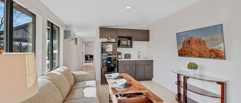 A modern living room with a sofa, a wooden coffee table, and a glass dining table. There is a kitchen area with cabinets and a refrigerator. A painting of a desert landscape is on the wall.