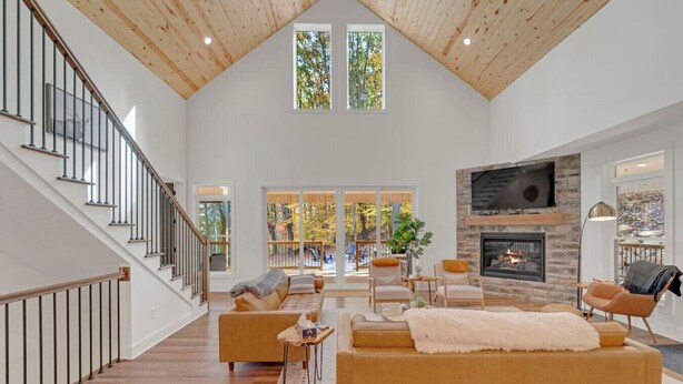 Un salón amplio con techo alto, chimenea, televisor grande y una escalera que lleva a un balcón con vistas a árboles.