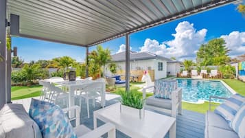 Un patio techado al aire libre con mobiliario blanco, una piscina y un césped.