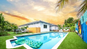 Une piscine avec des bouées gonflables, une clôture en bois et un bâtiment blanc doté d’un toit recouvert de tuiles.