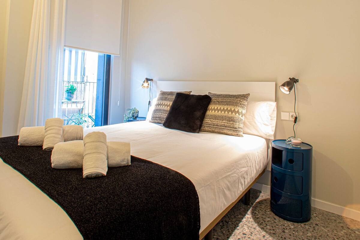 A bedroom with a bed, pillows, a nightstand, a lamp, and a window with curtains.