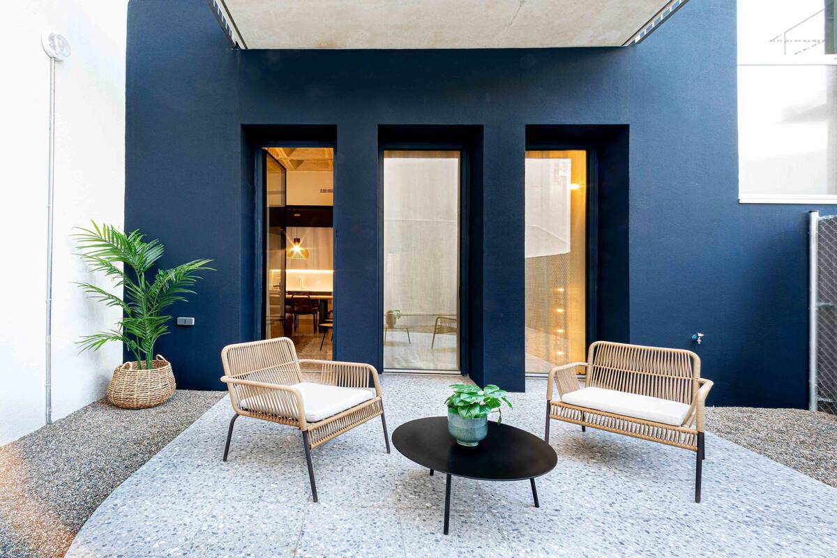 A modern outdoor seating area with wicker chairs and a central table, a dark blue wall, and a glass door leading to an interior space.