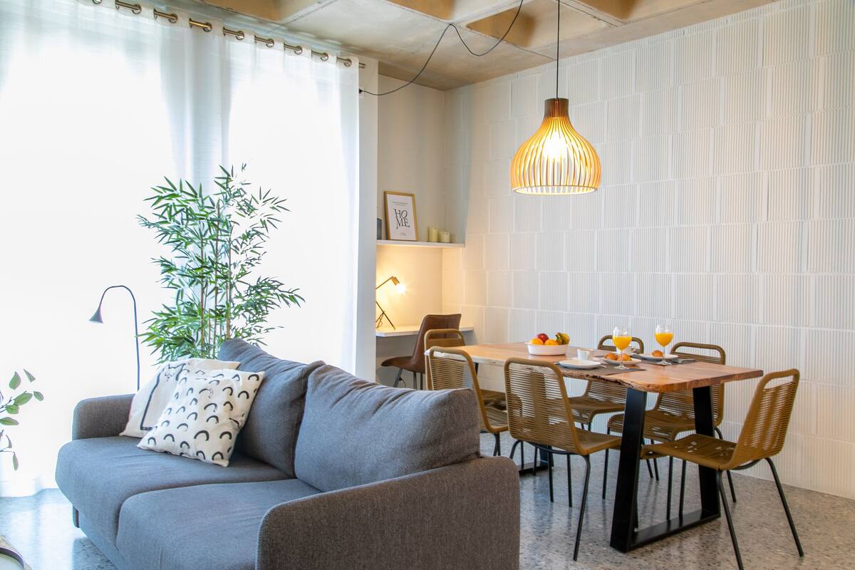 A modern living room with a grey sofa, a dining table with chairs, a hanging pendant light, and a bamboo plant.