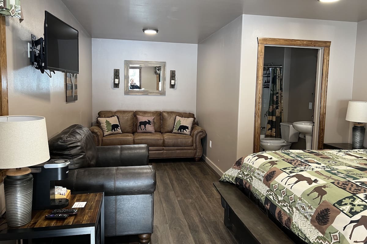 A hotel room with a sofa, a bed with patterned bedding, a TV mounted on the wall, a mirror, and a bathroom visible through an open door.