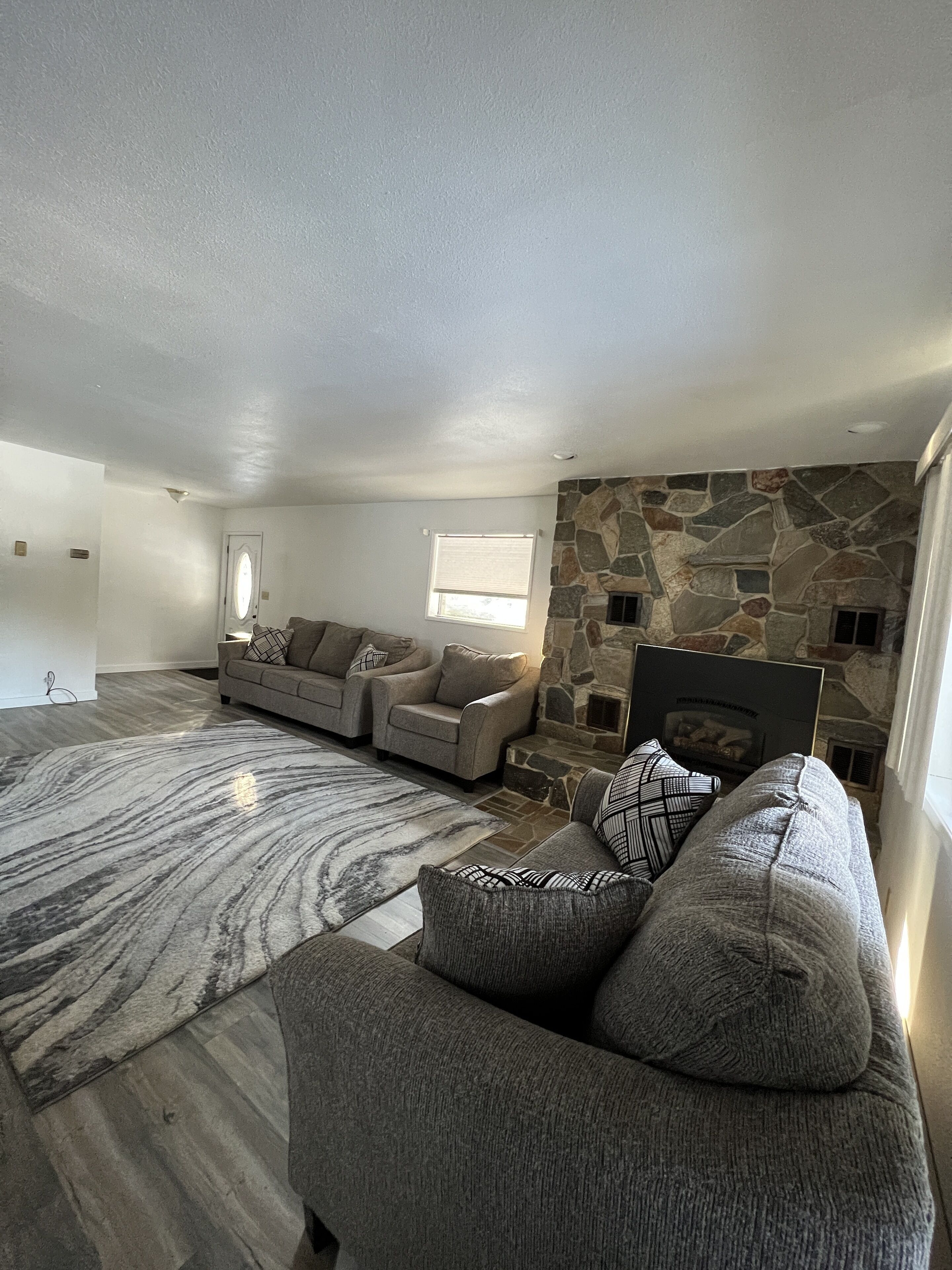 A spacious living room with a stone fireplace, two grey sofas, a patterned rug, and a large window.