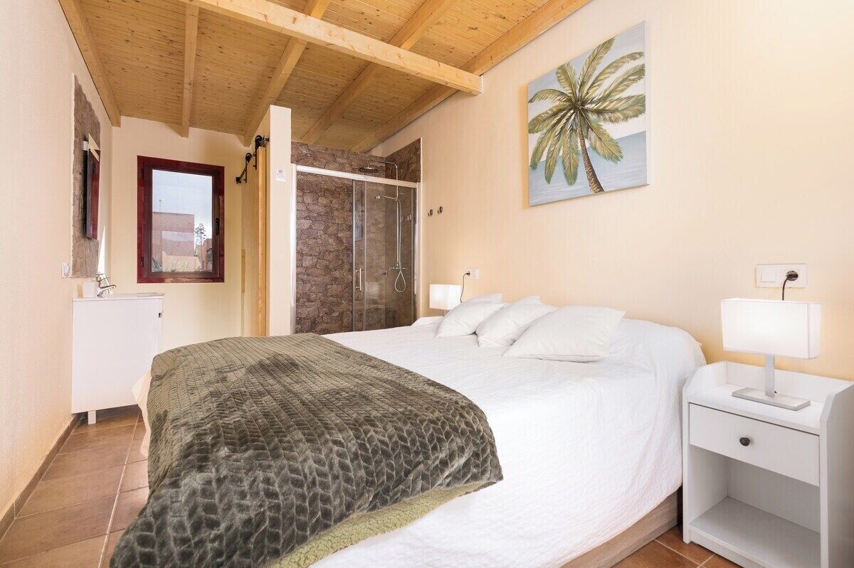Un dormitorio con una cama grande, una mesita de noche, una lámpara, un cuadro de una palmera y una zona de ducha.