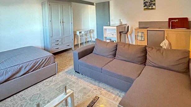 A modern living room with a sofa, a glass coffee table, and a small TV. There is a kitchenette area with a fridge and a microwave.