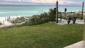 A grassy area with a wooden bench and a small table, overlooking a beach with a wooden fence and a clear view of the ocean.