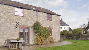 A stone building with a wooden door and windows, a small patio with a table and chairs, and a grassy yard. - 3 bedroom accommodation in North Wootton, near Wells (North Wootton, near Wells)