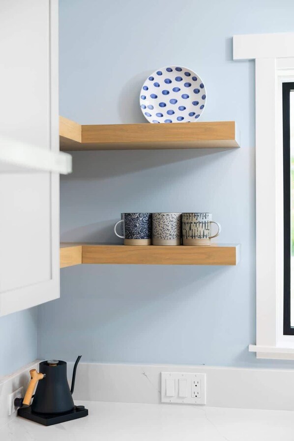 Une cuisine avec des meubles blancs, un mur bleu et des étagères en bois sur lesquelles sont posées une assiette et trois tasses.