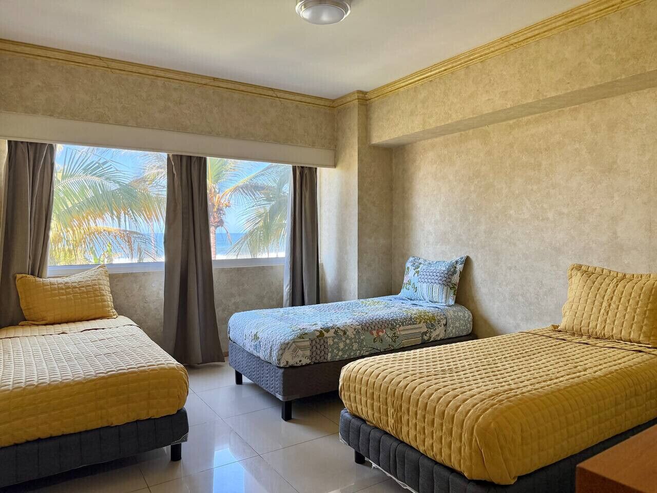 Une chambre d’hôtel avec trois lits, une grande fenêtre avec des rideaux et une vue sur des palmiers.
