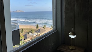 Habitación con un ventanal grande que da a una playa y al océano, una pequeña mesa con una lámpara, y vistas a una zona turística.