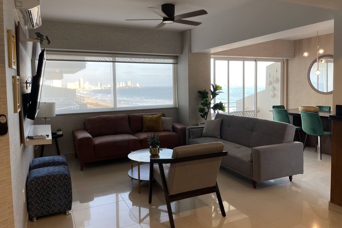 A modern living room with a sofa, armchair, and a small table. There is a large window with a view of a city skyline.