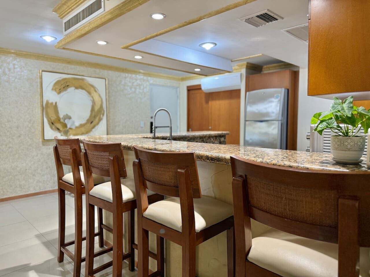 A kitchen with a granite countertop, wooden chairs, a stainless steel refrigerator, and a potted plant.