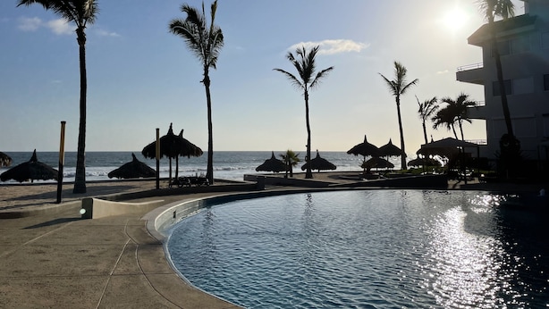 Vista de la piscina con palmeras, una playa y un edificio al fondo.