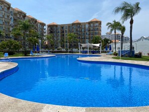 A large, clear swimming pool surrounded by lounge chairs and umbrellas, with multi-story buildings in the background. - Puesta del Sol (Mazatlán)