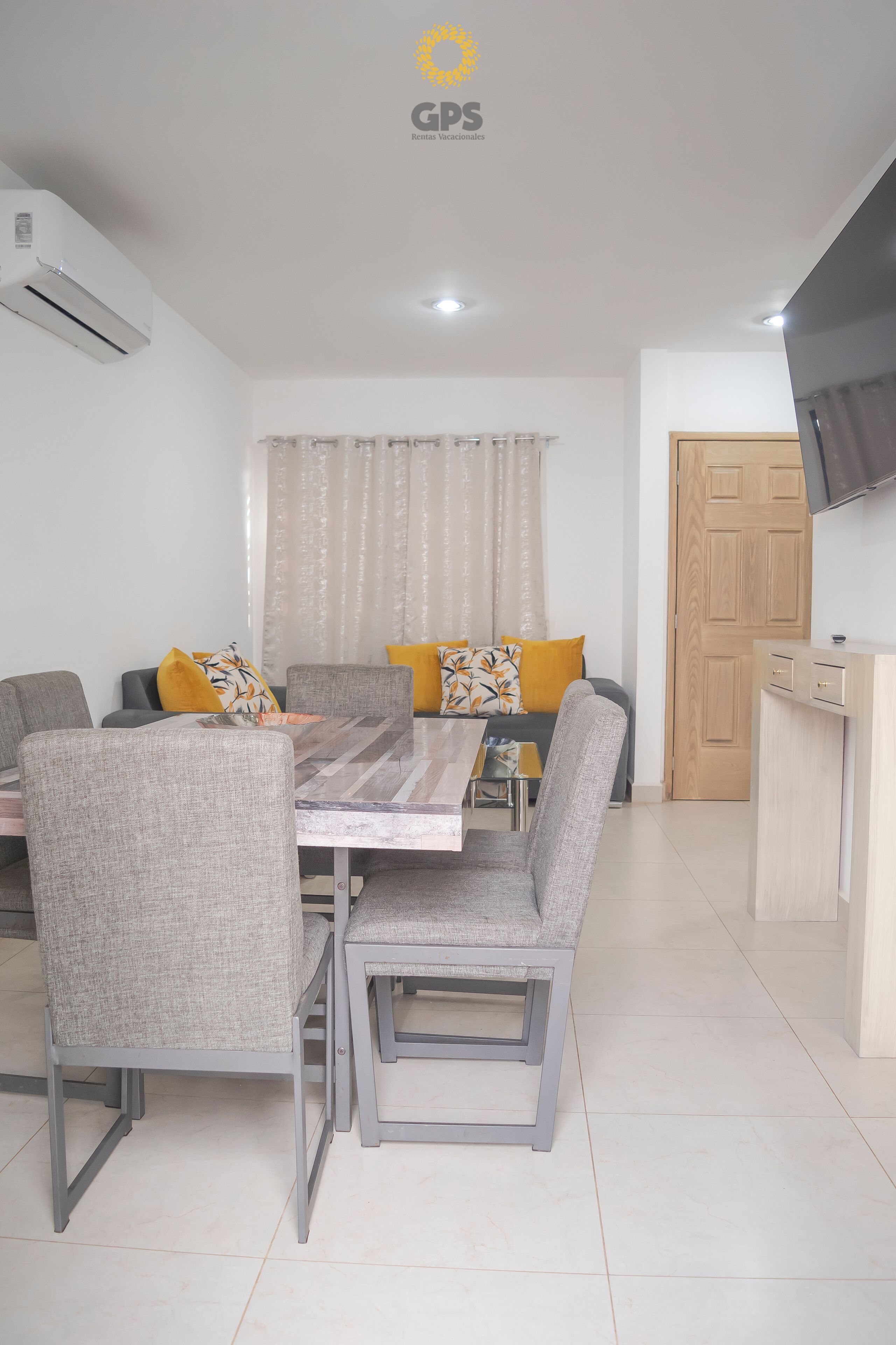 Una sala moderna con una mesa de comedor, sillas tapizadas en gris, un sofá con cojines amarillos, un televisor montado en la pared y una puerta de madera.