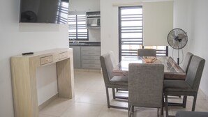 A modern dining area with a wooden table and grey upholstered chairs, a flat-screen TV mounted on the wall, and a standing fan. - Azul Marino (Mazatlán)