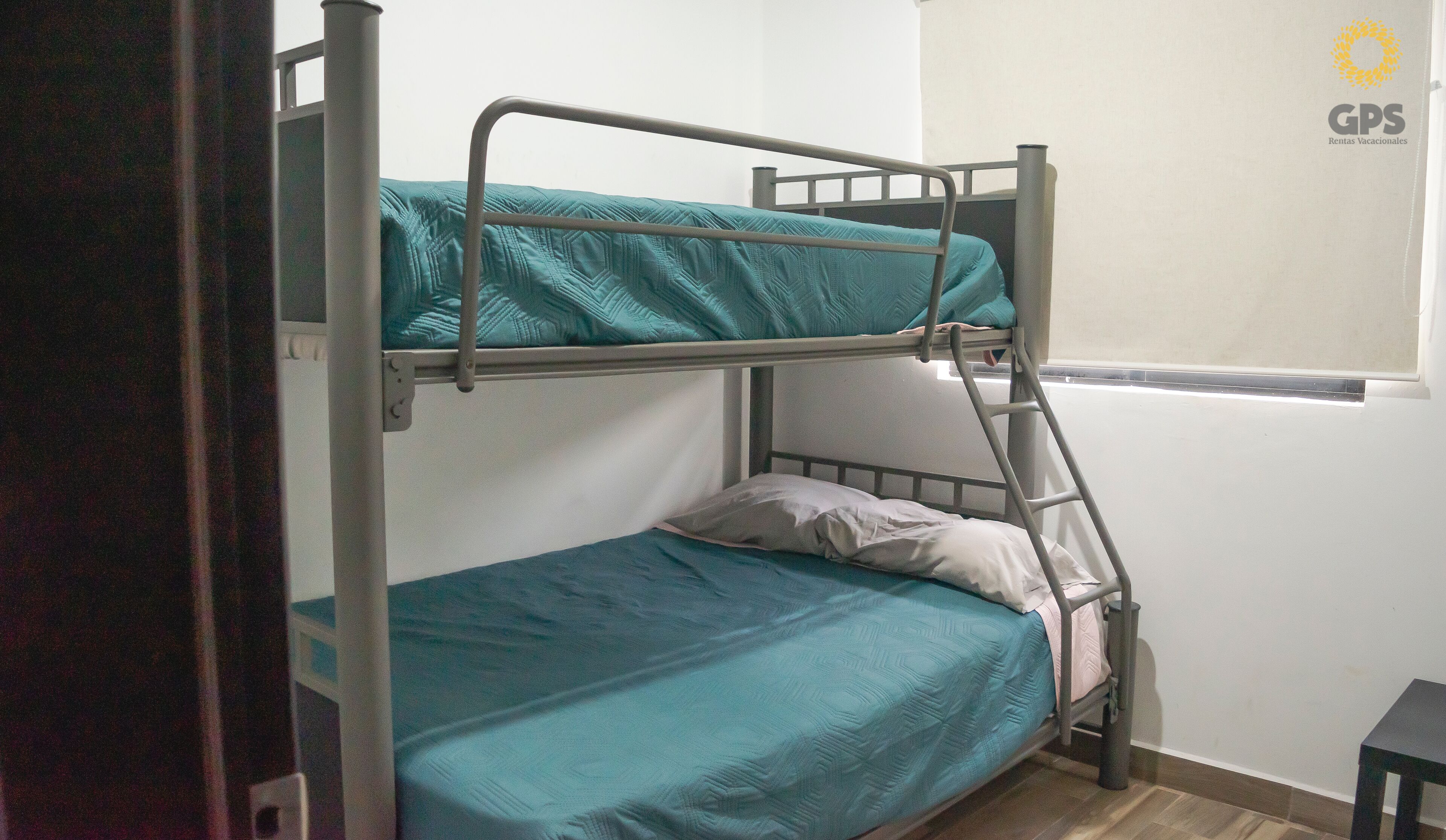 A bunk bed with a ladder, a window with blinds, and a small table.