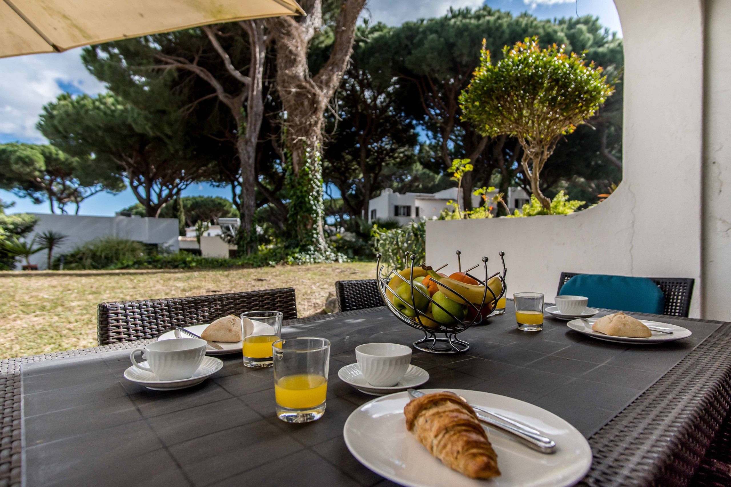 An outdoor dining area with wicker chairs, a table set with plates, glasses, and a fruit bowl.