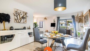 Una cocina moderna con armarios blancos, una mesa de comedor de vidrio y sillas grises. Cuenta con un amplio ventanal con cortinas, un televisor de pantalla plana y un elemento decorativo en la pared.